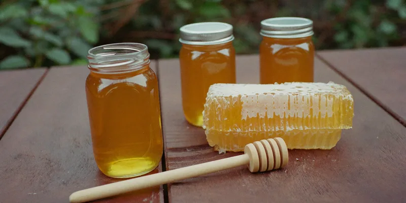 Plain glass jars of honey with honeycomb and a wooden dipper