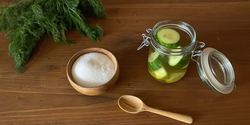 Bowl of white granules beside a jar of cucumber pickles and dill